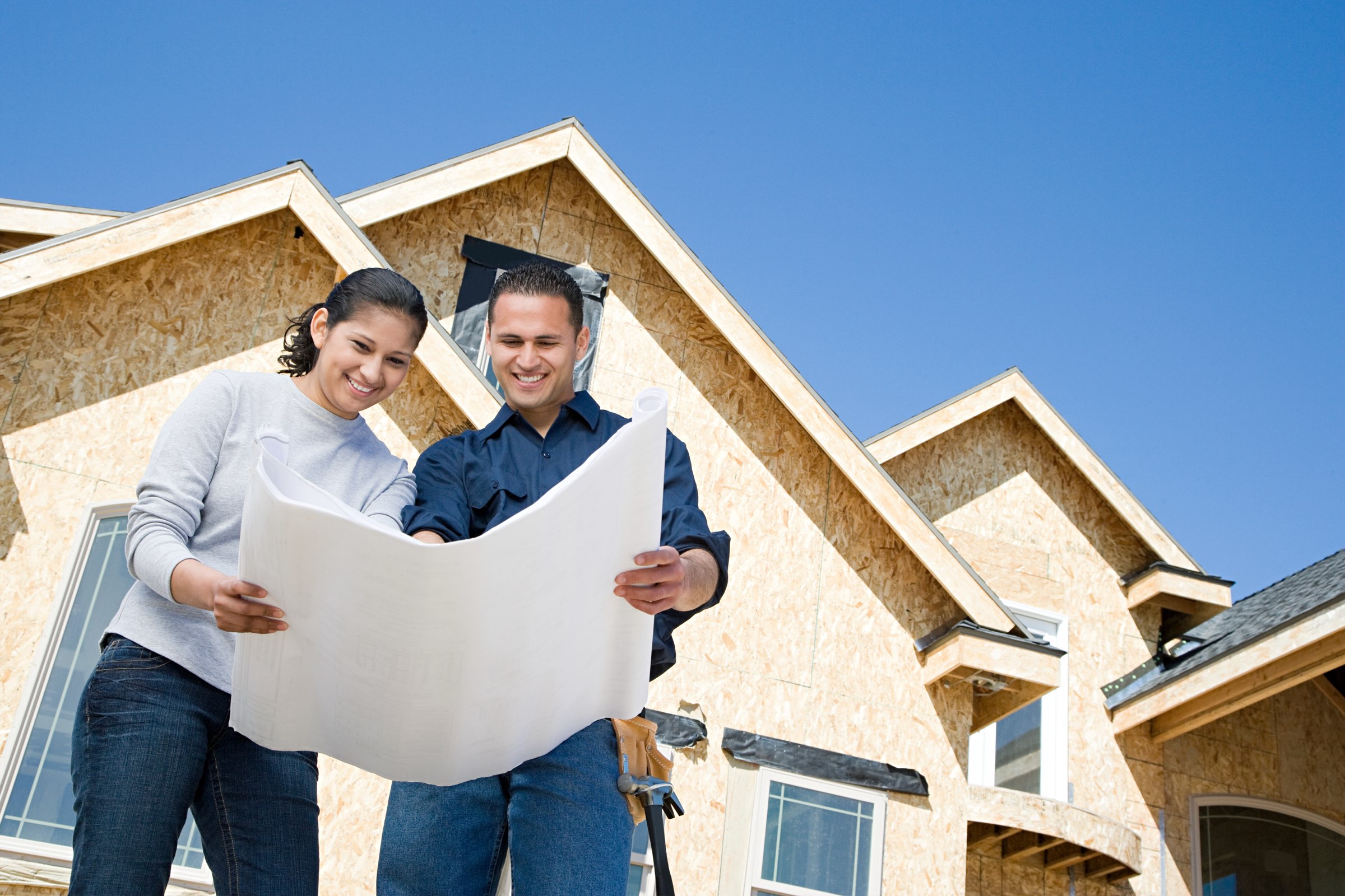 Relocation buyers reviewing house plans with a builder in front of a framed, sheathed new-construction spec home in a Johnson County, Kansas subdivision