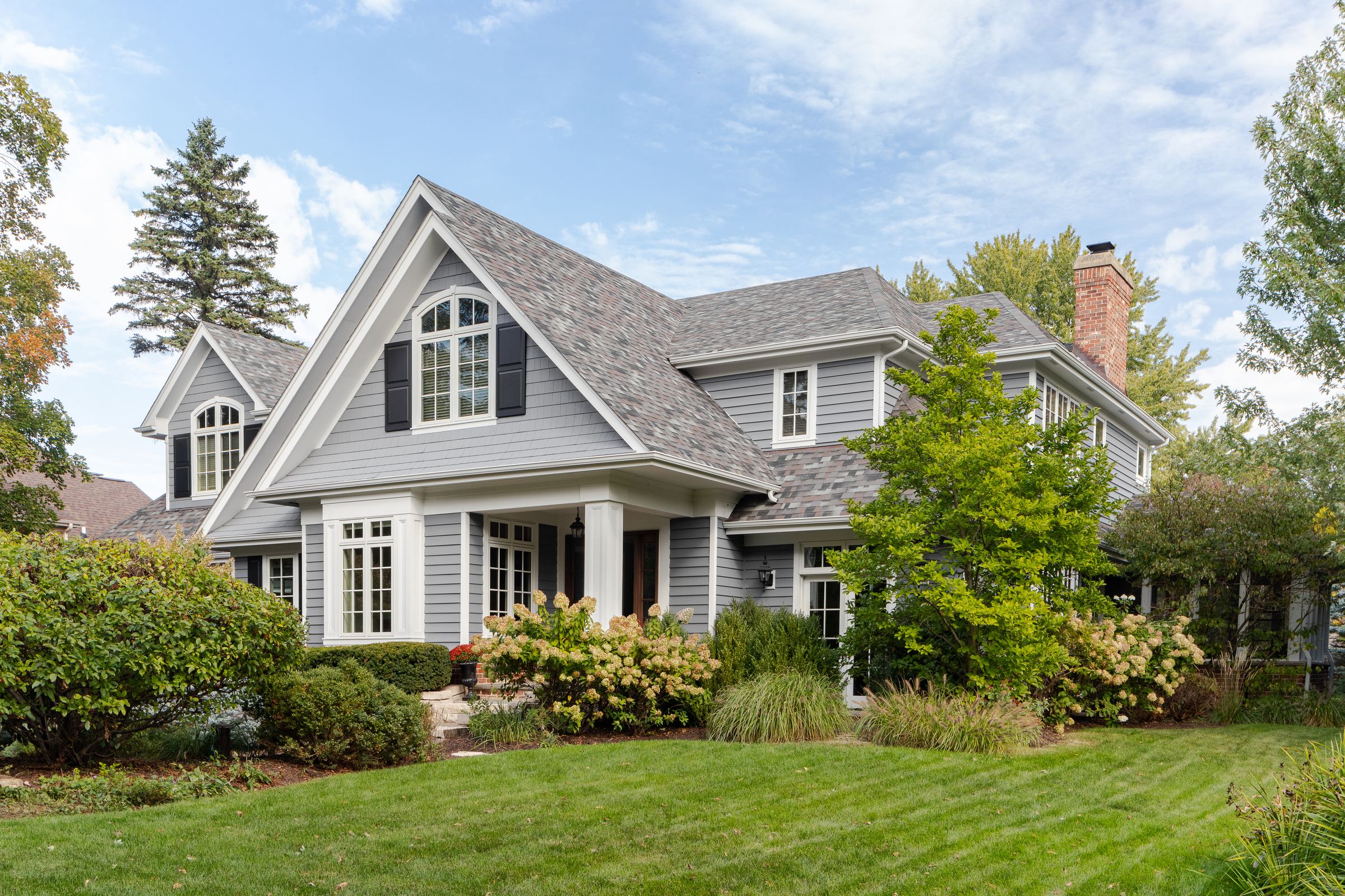 Established two-story gray shingle-and-lap-siding home with black shutters, an arched gable window, a covered front porch, and mature hydrangea landscaping on a manicured lawn in an Overland Park, KS neighborhood