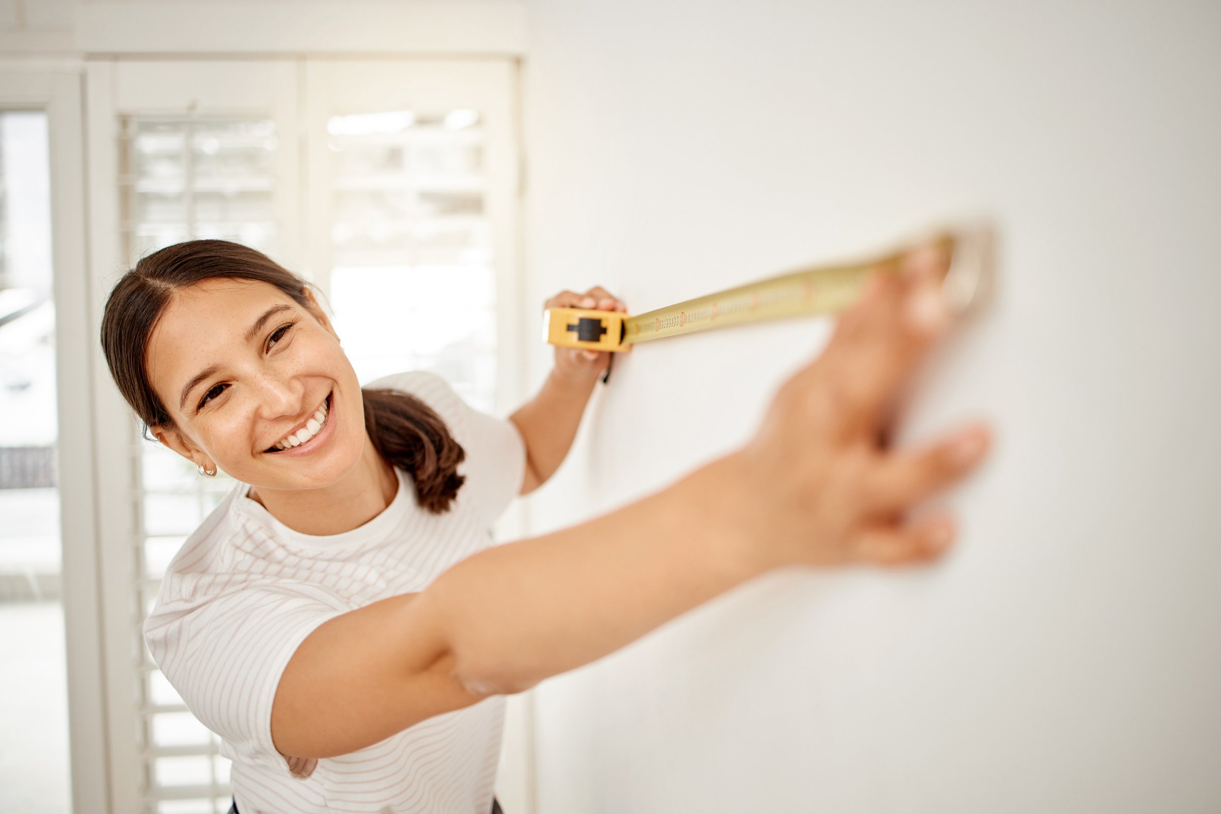A smiling woman in a white t-shirt stretches a yellow tape measure across a bright white interior wall to measure a room, in a sunlit space with plantation-shutter windows — illustrating how a home's price per square foot is measured